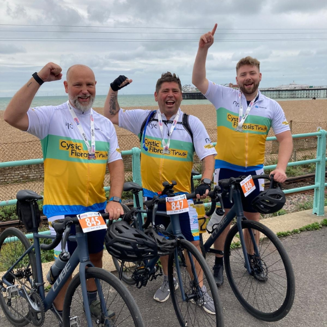 3 cyclists cheering and wearing cystic fibrosis jerseys in front of the beach