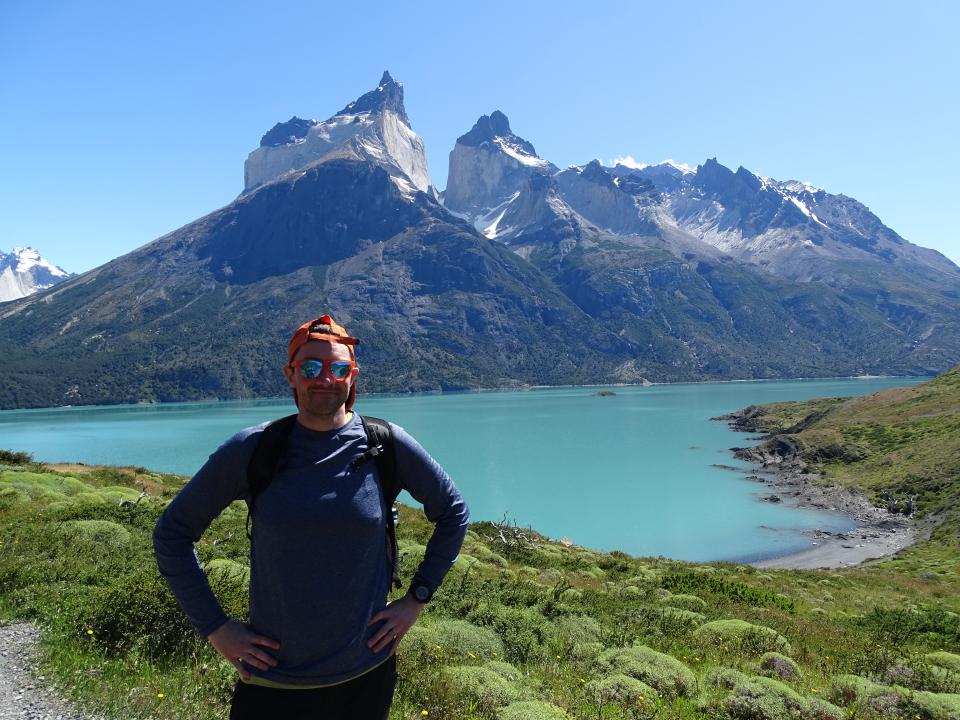 Josh stands with his hands on his hips, on holiday in front of a mountain and very blue water.