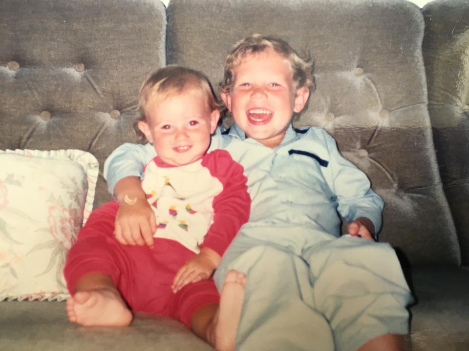 Mark is sat on the sofa, with his arm around his younger brother. They are both very young and smiling at the camera.