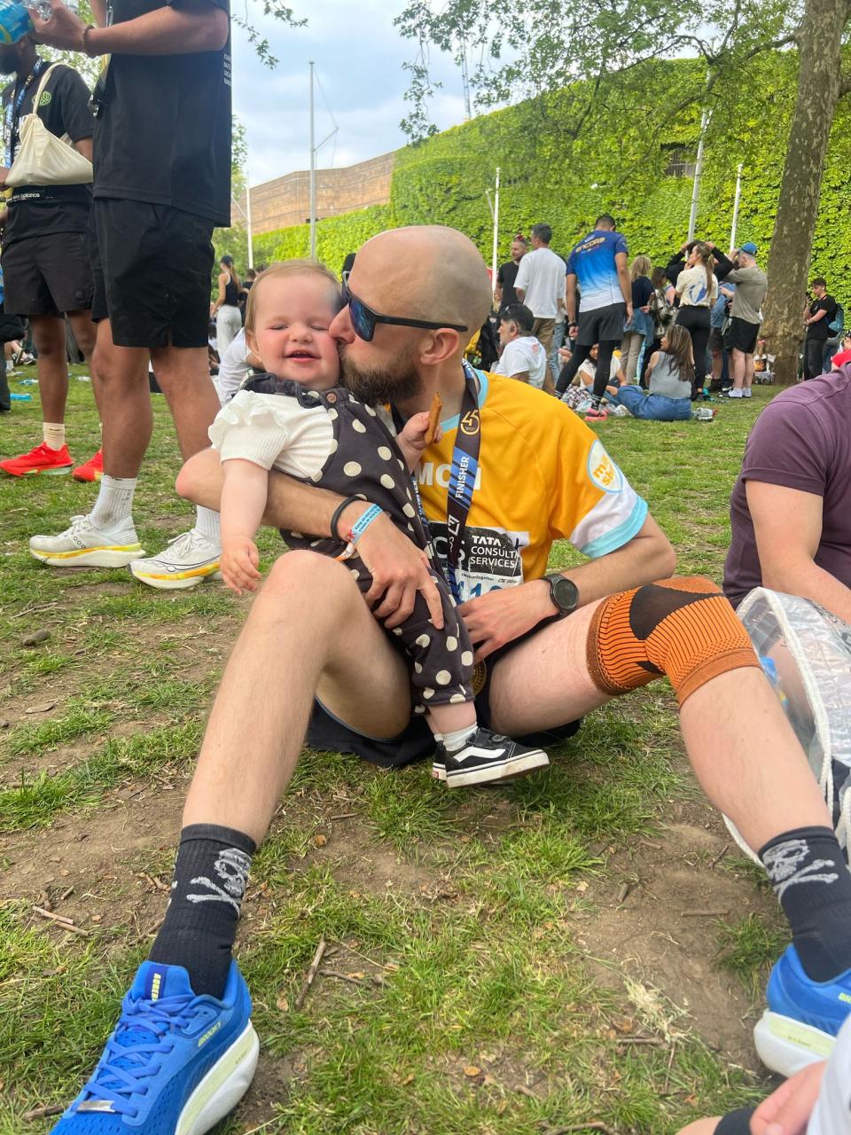 Simon and Ava. Simon sits on the grass in his running shirt, kissing daughter Ava on the cheek.