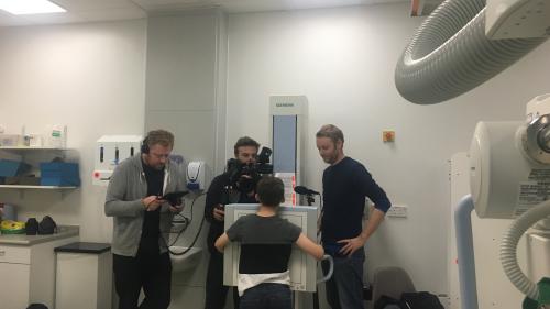 In a hospital a little boy is being scanned by a machine and surrounded by people with video recording equipment.
