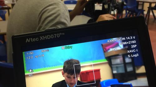 A screen with a little boy doing some work and in the background a man with a camera filiming the boy. 