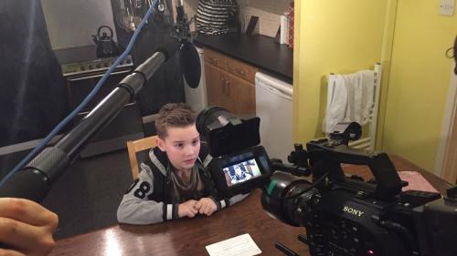 A video camera facing a little boy who is being interviewed sitting at a table in a kitchen. 