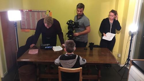 A little boy sitting at a wooden table facing a man with a video recorder, another man looking at a screen on the table and a lady with some paper in her hands.