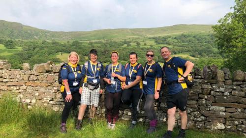 Six adults in walking gear, leaning against a stone wall with rolling hills behind them