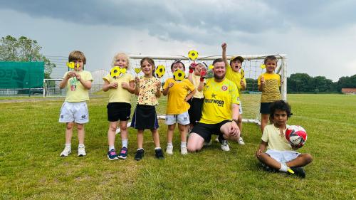 Eight young children and an adult male all wearing yellow on a football pitch, holding yellow heart and football placards