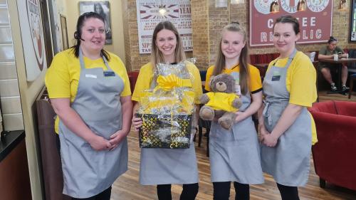 Four women wearing yellow t-shirts underneath barista aprons, holding a gift basket and teddy bear
