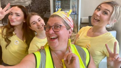 Group of four women posing, wearing yellow. One in a Hi Vis jacket. In a dental surgery.