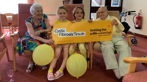 Two grandparents flanking two grandchildren, all wearing yellow and holding a Cystic Fibrosis Trust banner, with balloons spread around.