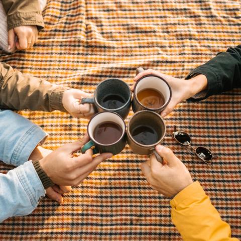 Cups of tea being clinked over a yellow checkered picnic blanket