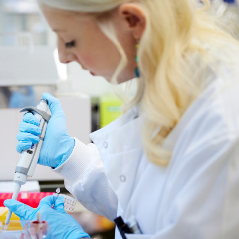 a blonde woman wearing a white coat in a lab pipettes solution into a tray