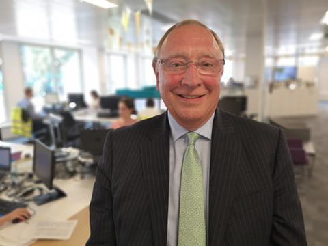 A middle aged man with wire-framed glasses, wearing a pin-stripe suit, light blue shirt and a pale green tie smiles at the camera in front of an office background.