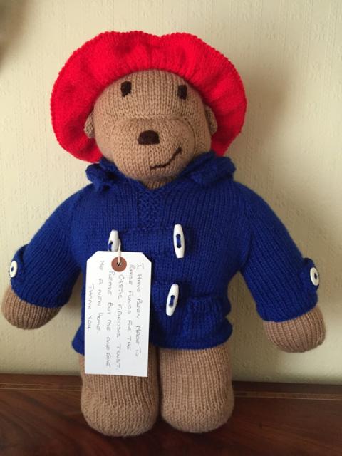 A knitted Paddington Bear toy. The bear is light brown, wearing a red hat and a dark blue coat with white buttons. He has a tag around his neck.