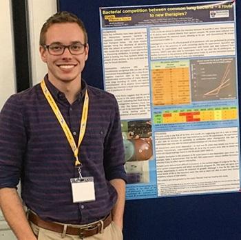 A young white man with fair hair, wearing black framed glasses, a checked shirt and a yellow lanyard, stands smiling in front of a poster