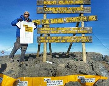 An Asian man with dark glasses wearing a grey beanie hat, blue jacket and grey trousers, stands on top of a mountain holding a white Cystic Fibrosis Trust branded tshirt. He is standing in front of a sign saying 'Mount Kilimanjaro'
