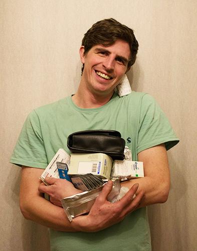 A white man with dark brown hair wearing a light green tshirt is holding lots of boxes of medication and smiling