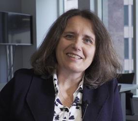 A middle aged Caucasian woman with medium length brown hair, wearing a blue jacket and white and blue shirt, smiles at the camera