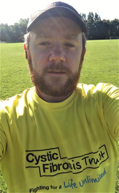 man with dark blonde beard wearing black cap, wearing yellow t-shirt that has 'Cystic Fibrosis Trust: fighting for a life unlimited' on chest