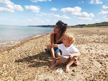 A white woman with brown hair and a young white boy with blonde hair sit on a pebble beach looking out at the sea.