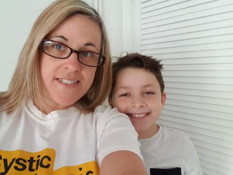 woman with blonde hair wearing glasses and white top with Cystic Fibrosis Trust logo, holding camera, small boy standing slightly behind with dark brown hair and white top, smiling