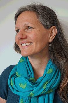 An image of Dr Samantha Phillips, who is Caucasian and has long salt and pepper hair and brown hair. Dr Phillips is wearing a blue scarf and earring and is smiling.