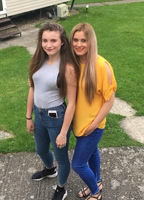A young girl with brown hair, wearing a grey tank top, blue jeans and black trainers stands next to a woman with blonde hair wearing a yellow top, blue jeans and sandals. Both of them are smiling at the camera and they are standing outside in a garden.