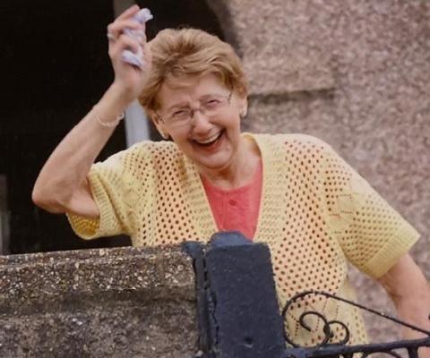 An older woman with light brown hair wearing a yellow cardigan and waving, standing behind a gate