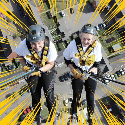 two women one slightly older in abseiling gear abseiling down a building, wearing Cystic Fibrosis Trust t-shirts and looking up