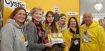 Six people stand in a line smiling at the camera; three women and one man. The woman in the middle holds a photo of a young child holding a sign saying #OrkambiNow. They are all wearing yellow or Trust-logo sweatshirts and they are standing in front of a sign with the Trust's logo on it and facts about CF on it.