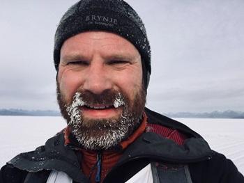 A white man with a brown beard wearing a hat smiling. He has frost in his beard.