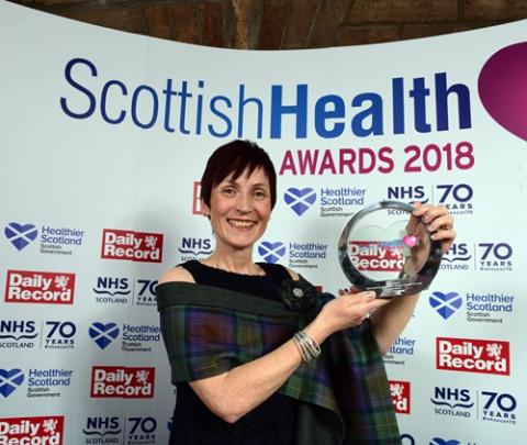 A white woman with cropped dark brown hair holds up a circular glass award in front of a board with 'Scottish Health Awards 2018' written on it. She is wearing a black sleaveless top with a tartan wrap over her shoulders.