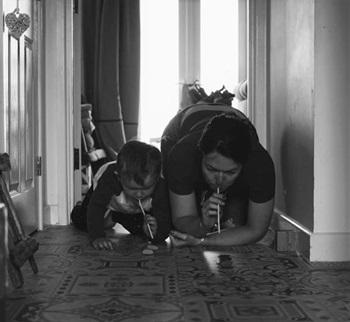 black and white photo of mum and son kneeling down and blowing something along the floor with straws