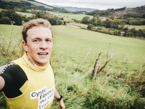 young white man with blonde hair wearing black running tshirt under a yellow Cystic Fibrosis Trust running vest, holding camera in selfie mode with field landscape background