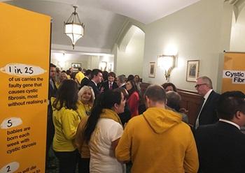 A large group of people mill around in a high-ceilinged white room. Most of them wear yellow, and there are is a sign giving statistics about cystic fibrosis.