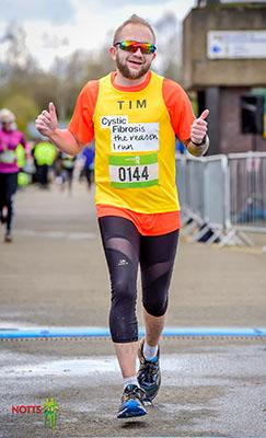 A man with a brown beard in a yellow running vest puts his thumbs up while he's running.