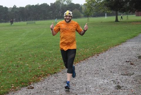A man with a beard in an orange t-shirt and a yellow bandanna runs through a park.