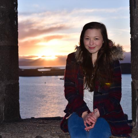 girl around 20's sitting on castle wall with sunset in background, long dark brown hair wearing maroon winter coat and blue jeans
