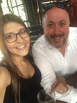chriswoman with long brown hair wearing glasses and blank tank top linking arms with man with sunglasses on head and beard wearing white collared shirt sat at a table with a drink
