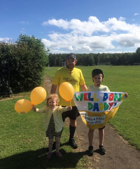man in running gear wearing navy cap and yellow t-shirt standing with young girl and boy both holding balloons and banners, standing on a football pitch