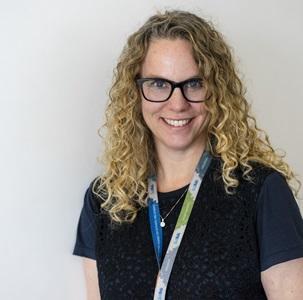 woman with blonde curly hair wearing glasses and a navy blue shirt with a lanyard 