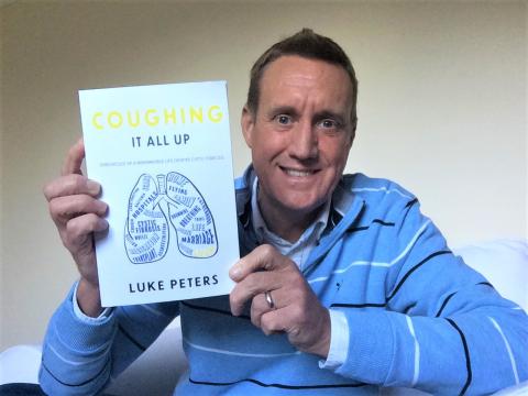 Man holding up a book called Coughing it all up by Luke Peters
