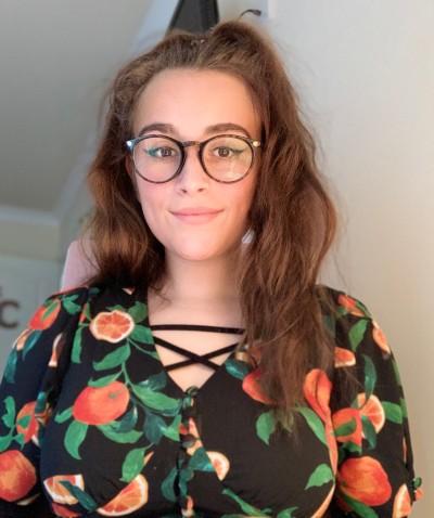 Young white woman with long hair, wearing glasses and a black dress with orange (the fruit) print
