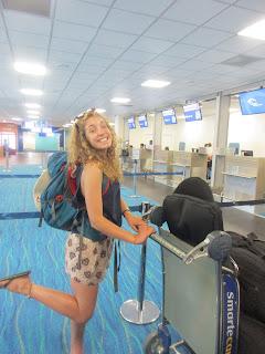 Nicole stands in an airport, smiling and pushing a luggage trolley