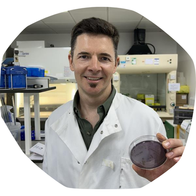Dr Luke Allsopp in his lab, wearing a white lab coat and holding a petri dish