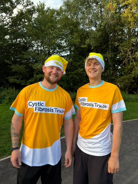 2 men smiling, wearing cystic fibrosis trust yellow tops with yellow Santa hats